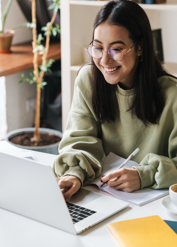 Smiling student with glasses makes notes in a notebook at a kitchen table.